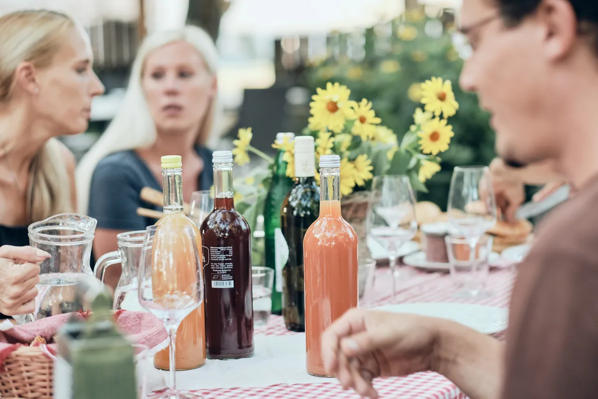 Gäste im Gespräch an einer gedeckten Tafel mit Sonnenblumen und Säften im Garten des Biohofs ADAMAH
