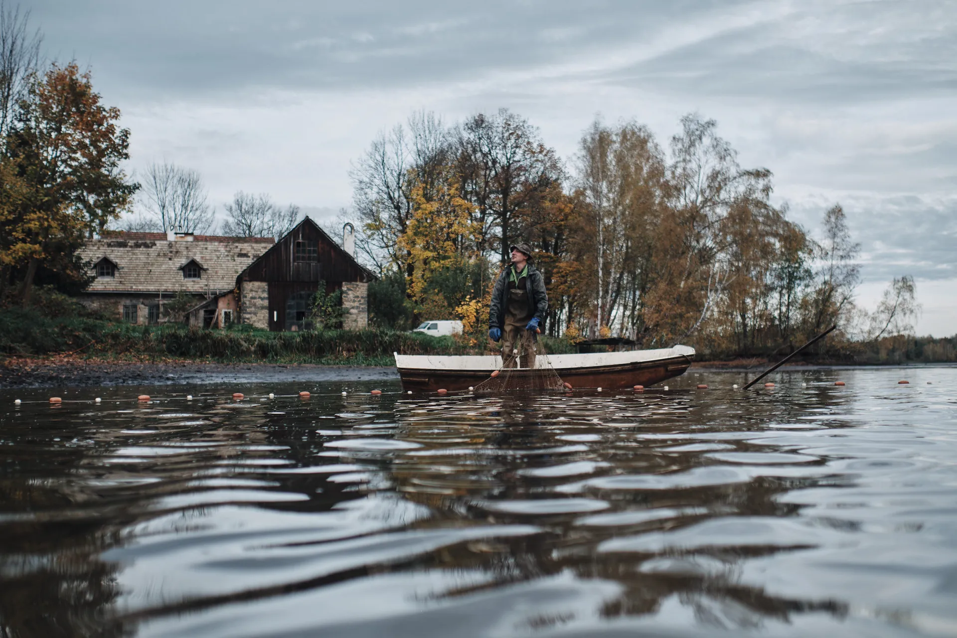 Ein Fischer holt vom Boot aus das Netz ein, am Ufer ein altes Teichhaus aus Stein und Holz.