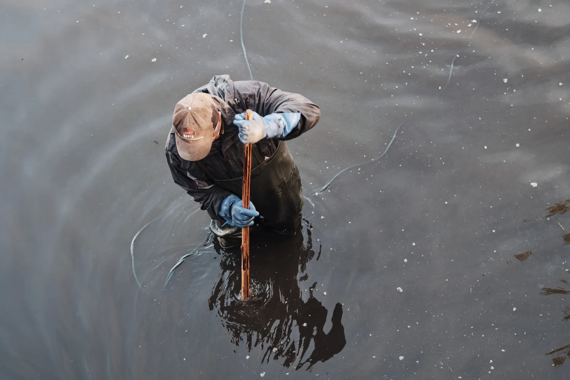 Von oben gesehen steht ein Fischer bis zu den Hüften im grauen Wasser und hält eine Stange.