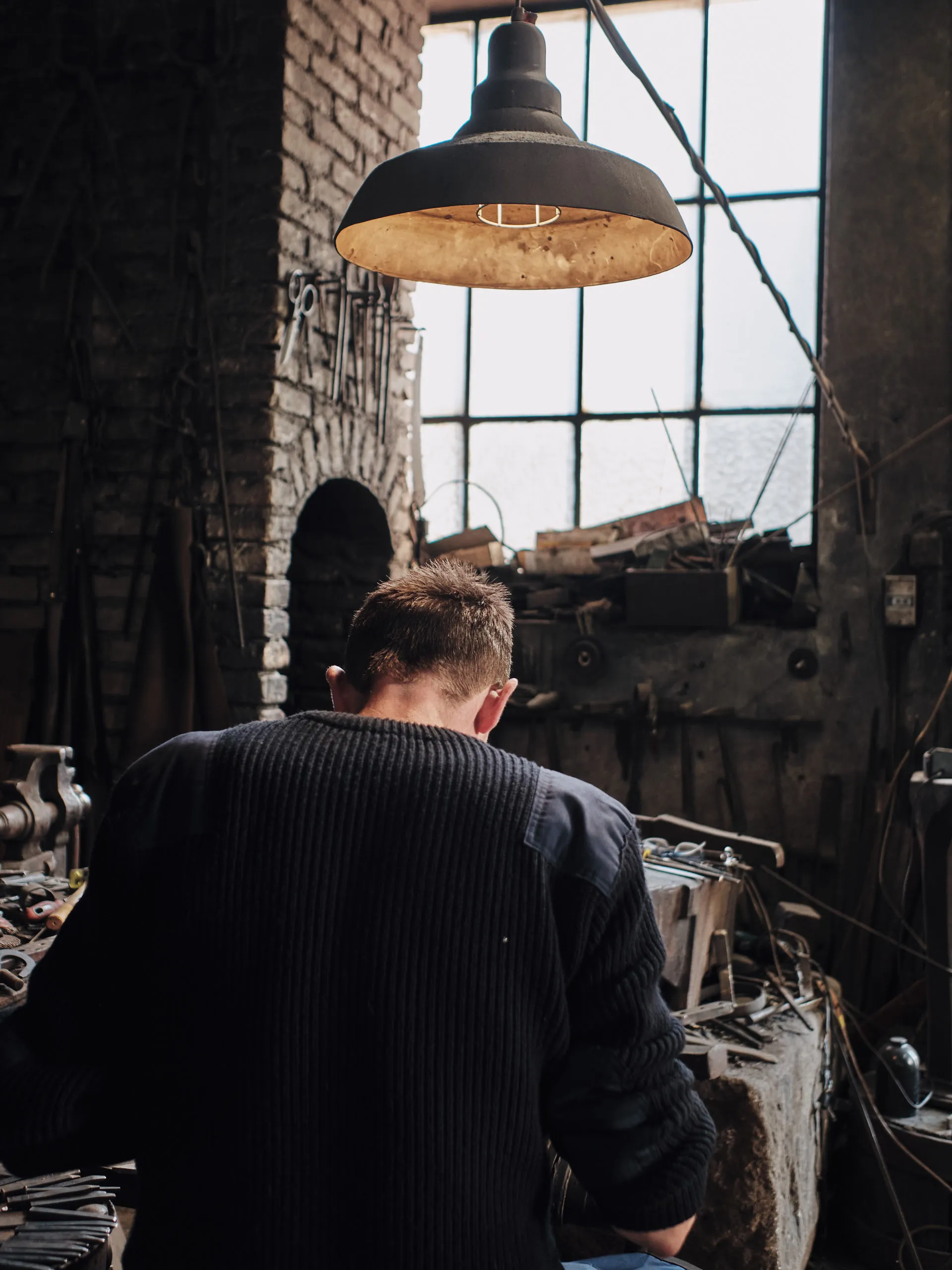 Blick von hinten in die alte Schmiedewerkstatt der Gebrüder Schmidberger in Molln, ein Schmied gebeugt über den Werktisch unter der Werkstattlampe.