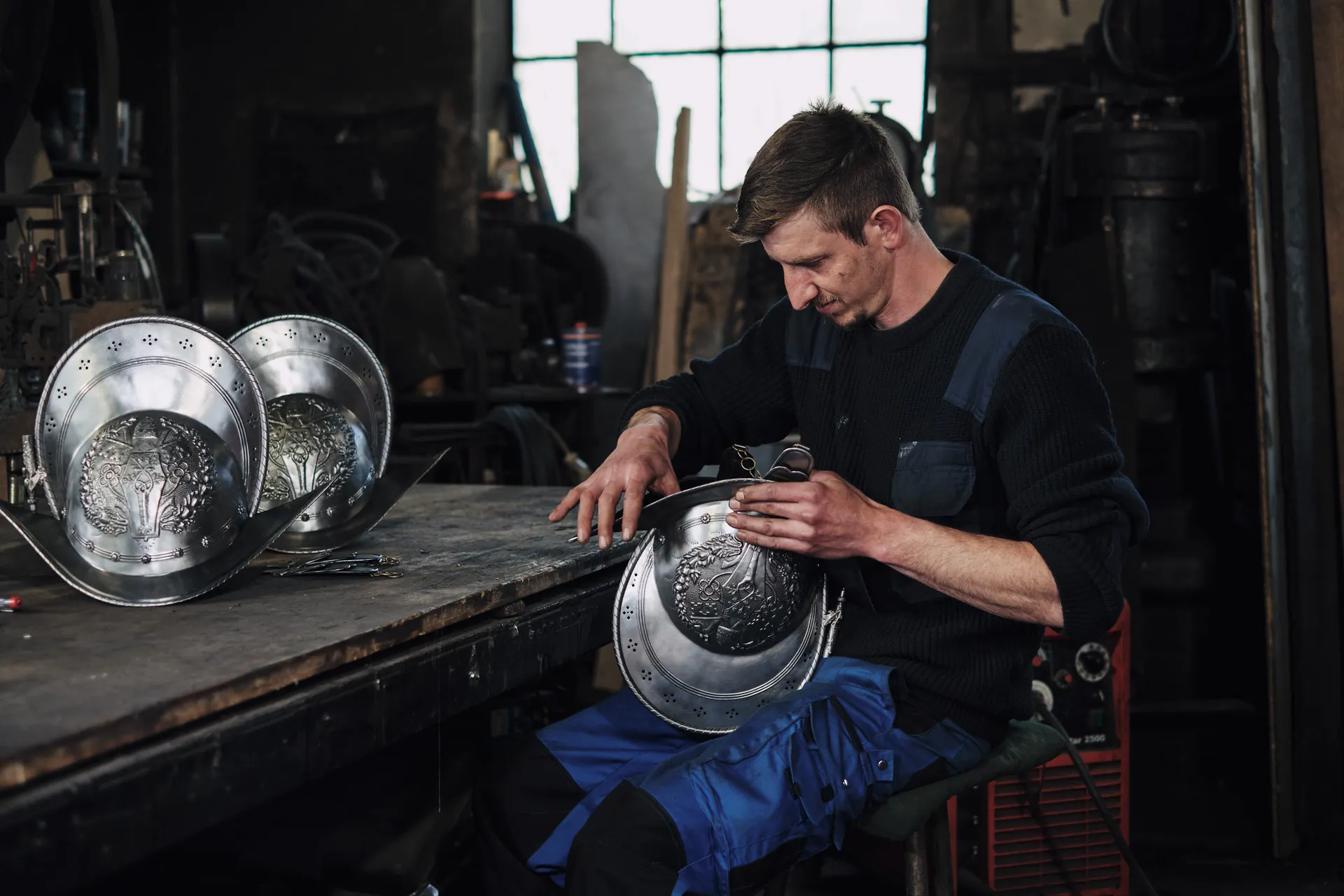 Ein Schmied vollendet einen verzierten Helm, daneben zwei fertige Helme der Schweizergarde auf dem Werktisch.