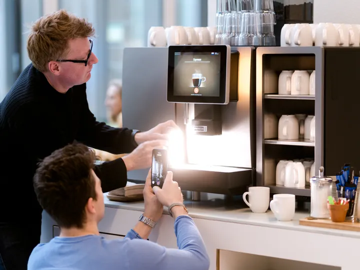 Wolfgang Lehner zeigt einem Mitarbeiter an der Kaffeemaschine, wie er die Szene mit dem Smartphone aufnimmt